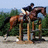 Pixelated image of a person riding a horse in mid-jump over an obstacle, set against a blurred background of greenery and sky.