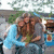 Three people embrace outdoors with a blue vehicle and greenery in the background, captured