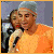 Pixelated image of a person holding a microphone, wearing an orange shirt and dark jacket against a blurred background.