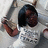 Pixelated static image of a person with dark hair and brown skin wearing a white shirt, holding an object near their face.