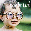 A child wearing large round glasses with brown frames and pink-tinted lenses, standing against a blurred green background with the word 'rockstar' in white text above the head.
