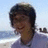 A person with short dark hair wearing a light purple shirt smiles against a beach background with water and sky.