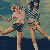 Two women in casual summer clothing pose dynamically against a blurred blue background, one in a pink shirt and green shorts, the other in a striped shirt and plaid shorts.