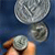 A hand holds a silver coin with a central hole against a blue background, with two additional coins visible above it.