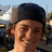 Pixelated close-up of a person with light brown hair wearing a dark blue cap, smiling against an outdoor background.