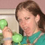 A person with brown hair wearing a teal shirt holds two green spherical objects near their face against a light background.