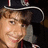 Pixelated image of a person wearing a dark hat with white trim and gray clothing, next to red pixelated text on black background.