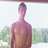 Pixelated static image of a shirtless figure with short dark hair standing against a blurred outdoor background with green and blue tones.