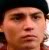 A pixelated close-up of a person's face with a dark cap, neutral expression, and blurred background.