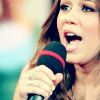 A close-up of a person holding a black microphone with a red stripe near the top, mouth open as if singing or speaking, long brown hair visible, blurred background.