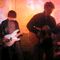 A band of four musicians plays electric guitars under warm orange lighting, with the scene shifting from left to right