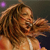 A pixelated figure with long brown hair holds a microphone, wearing a light-colored top against a dark background with purple accents.