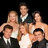 Six people in formal attire pose together in a group, with three standing behind three seated individuals against a dark background.