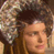 Pixelated static image of a person with brown hair and light skin tone, wearing a large dark headpiece or hat with textured patterns on the left side.