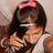 A pixelated image of a person with long dark hair wearing a pink bow and white top, next to a pink background with repeated purple text.