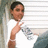 Pixelated image of a person with short dark hair wearing white shirt and blue pants, making peace sign gesture against striped background.