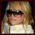 Pixelated static image of a person with long blonde hair wearing dark sunglasses and a white top against a dark background.
