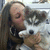 A person with long brown hair holds a small white and gray dog with dark eyes and nose, positioned close to the face.