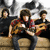 Three people sit on a couch holding guitars, with the central figure's guitar slightly shifted between frames.