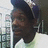Pixelated static image of a person with dark hair and brown skin wearing a dark blue top against a blurred background with light and dark areas.