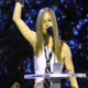 Four of a person with long brown hair singing into a microphone at a podium, wearing a white sleeveless top and holding an award
