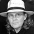 Black-and-white pixelated image of a person wearing a wide-brimmed hat with a dark band, face partially visible with neutral expression.