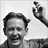 Black-and-white pixelated image of a person with short dark hair and a mustache, smiling slightly while raising their right hand in a gesture.