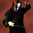 Pixelated figure in black hooded garment with white mask-like face, holding glowing object against brown background.