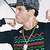 Pixelated figure in dark jacket with colorful stripes, wearing patterned cap and glasses, pointing right against light background.