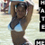 A woman in a light blue bikini top and dark bottoms poses with arms raised above her head, set against a blurred beach background.