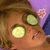 A person with light brown hair lies back with two cucumber slices over their eyes, set against a purple background with pink floral patterns.