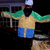 Pixelated figure in green long sleeves, beige torso, blue shorts, arms extended horizontally against dark background with faint structure.