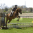 A pixelated static image shows a rider on horseback mid-jump over an obstacle in a grassy field with blurred background structures.