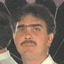 A person with dark hair and a mustache wears a white shirt against a dark background with subtle light patterns.