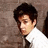 Pixelated static image of a person with dark hair wearing a white shirt and dark tie against a brown textured background.