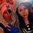 Two figures with light skin tones pose closely together; one wears a red head covering and blue garment, the other has dark hair and black clothing.