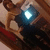 A pixelated image of a person in a dark outfit holding a glowing blue object, with a red and brown background.