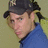Pixelated close-up of a person wearing dark cap and blue shirt against muted green background.