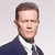 Pixelated portrait of a person with short brown hair wearing a dark blue suit jacket over a white collared shirt against a light gray background.