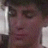 A pixelated close-up of a person's face with dark hair, visible eyes, nose, and mouth, set against a dark background with light vertical elements on the sides.