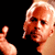 Pixelated static image of a person with light hair and visible facial features against black background, showing partial upper body and hand gesture.