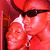 Two people in close proximity, one wearing dark sunglasses and a red garment, the other with a white headband and light-colored clothing, against a red background.