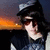 Pixelated image of a person with dark hair wearing sunglasses and a cap, holding an object near their face against a sunset background.