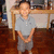 A child in a gray polo shirt and gray shorts stands indoors with hands clasped, smiling at the camera.