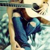 A person in blue ripped jeans and a dark top sits on a patterned blanket holding a light-colored acoustic guitar, with a dog partially visible behind them.