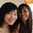Two people with long dark hair stand close together, smiling at the camera against a light background.