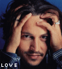 A person with long dark hair and a mustache holds both hands to their head against a blue background, wearing a checkered shirt.