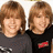 Two people with light brown hair stand side-by-side against a white background, while adjacent blue displays pink pixelated text
