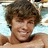 Pixelated close-up of a person with curly brown hair, light skin tone, and visible neck against a blurred background.