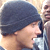 Pixelated static image of a person wearing a dark beanie and light-colored shirt, with partial view of another figure in the background.